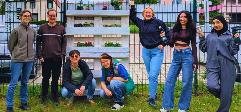 Sieben Personen stehen an einem Zaun und blicken in die Kamera