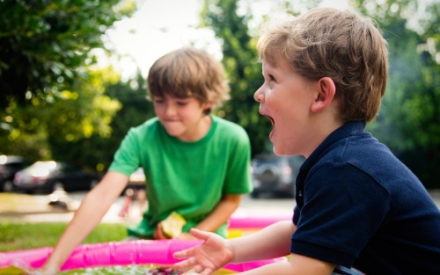 Zwei Jungen spielen mit Wasser in einem rosa Planschbecken. Der Kleinere von beiden freut sich und lacht.