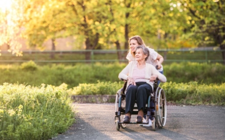 eine junge Frau schiebt eine Seniorin im Rollstuhl &uuml;ber einen Weg im Gr&uuml;nen