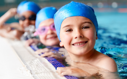 Am Rand eines Schwimmbeckens sind drei Kinder zu sehen. Sie halten sich am Beckenrand fest, tragen blaue Schwimmkappen und und l&auml;cheln in die Kamera.