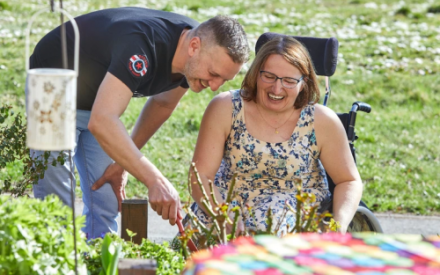 Ein Mann und eine Frau im Rollstuhl beugen sich &uuml;ber die Zweige einer zur&uuml;ckgeschnittenen Rose. Der Mann h&auml;lt ein Gartenwerkzeug in der rechten Hand.