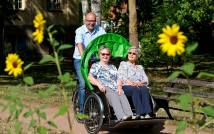 Zwei &auml;ltere Frauen sitzen in einer Rikscha mit gr&uuml;nem Dach. Die Rikscha wird von einem Mann gefahren und gelenkt. Im Vordergrund sind gelbe Sonnenblumen zu sehen.