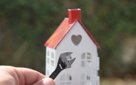 Im Hintergrund ein kleines wei&szlig;es Modellhaus mit rotem Spitzdach, unter dem Giebel ein Herz; davor ist eine Hand zu sehen, die eine Zange h&auml;lt