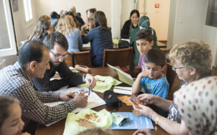In einem Raum sitzen an zwei Tischen mit Familien mit Kindern. Eine Frau spielt mit einem Jungen, ein Mann erkl&auml;rt einem anderen etwas in einem Buch