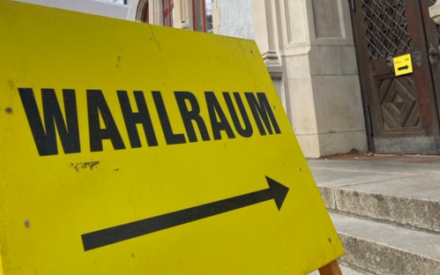 In Nahaufnahme ist ein leuchtend gelbes Schild zu sehen, auf dem in schwarzen Gro&szlig;buchstaben "WAHLRAUM" steht. Der Pfeil darunter zeigt nach rechts auf eine T&uuml;r, durch die es zum Wahlraum geht.