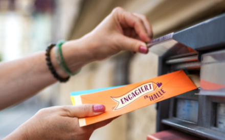 Vor einer Wand mit Briefk&auml;sten ist eine Frauenhand mit lackierten N&auml;geln zu sehen. Die Hand steckt gerade Post in einem Briefschlitz. Auf dem Briefumschlag ist das Logo "Engagiert in Halle" zu sehen.