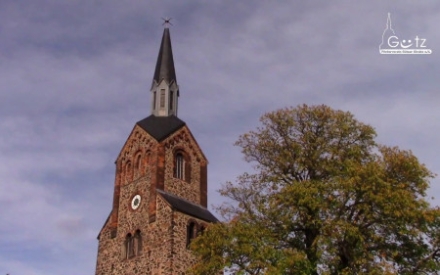vor einem blauen Himmel mit wenigen Wolken ist der Turm der G&uuml;tzer Kirche zu sehen, rechts davon ein gro&szlig;er Baum
