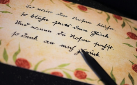 Auf einem Blatt Papier, das mit Blumen verziert ist, ist ein Vers in altdeutscher Schrift zu lesen.