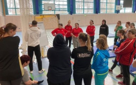 Eine Sportgruppe steht im Halbkreis um den Trainer, der auf ein Flipchart-Papier schreibt und etwas erkl&auml;rt.