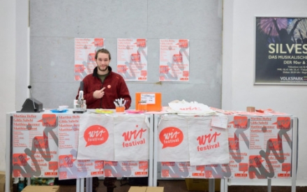 An einem Infostand vom WIR-Festival steht ein junger Mann. Vor ihm auf dem Tisch liegen Poster, Flyer und Stoffbeutel mit der f&uuml;r das Festival typischen Sprechblase in der Farbe orange.