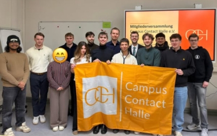 Junge Menschen stehen in zwei Reihen und halten eine Fahne in den Farben Orange und Wei&szlig; mit dem Schriftzug Campus Contact Halle e. V. in den H&auml;nden.
