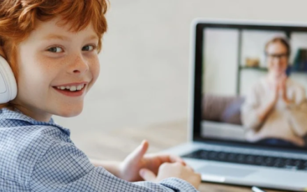 Ein Junge sitzt mit Kopfh&ouml;rern vor einem Laptop und l&auml;chelt in die Kamera. Auf dem Bildschirm ist eine l&auml;chelnde Frau mit Brille zu sehen.