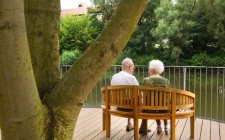 Eine Seniorin und ein Senior sitzen auf einer Bank am Fluss und schauen auf das Wasser. Beide sind von hinten zu sehen.