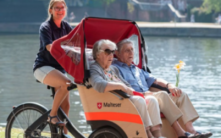 Zwei Senior:innen, eine Frau und ein Mann, sitzen in einer Rikscha, die an einem gew&auml;sser vorbeif&auml;hrt. Die Fahrerin der Rikscha l&auml;chelt in die Kamera.