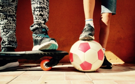 Auf dem Foto sind zwei Paar Beine vor einer rot gestrichenen Wand zu sehen. Ein Paar Beine in schwarz-wei&szlig; gemusterter Hose steht auf einem Skateboard. Das andere Paar Beine stejt mit dem rechten Fu&szlig; auf einem wei&szlig;-roten Fu&szlig;ball.