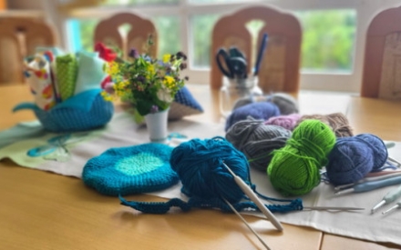 Auf einem Holztisch liegen mehrere Wollkn&auml;uel in Blau und Gr&uuml;n sowie mehrere H&auml;kelnadeln. Dahinter steht eine kleine Vase mit Blumen sowie ein Glas mit Scheren und Stiften.
