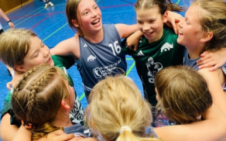 Eine Gruppe jugendlicher Sportlerinnen bildet einen Kreis und umarmt sich. Sie freuen sich und lachen. Die Gruppe ist von oben zu sehen und steht auf dem blauen Fu&szlig;boden einer Turnhalle. Auf der Sportkleidung ist das Logo der Basketball-Sektion USV Rhinos, ein Nashorn, zu sehen.
