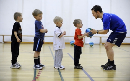 vier Kinder imVorschulalter stehen in der Turnhalle vor ihrem Trainer, der dem ersten Jungen einen Ball in die Hand gibt