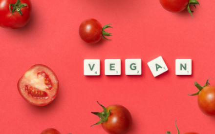 Auf rotem Hintergrund liegen einige Tomaten verteilt, manche sind aufgeschnitten - In der Mitte liegt das Wort "vegan" mit Buchstabenplatten gebildet, wie im Spiel Scrabble.