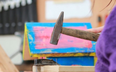 In Nahaufnahme ist ein kleiner Hammer zu sehen, der von einer Kinderhand gehalten wird und mit dem ein Nagel ins Holz geklopft wird. Im Hintergrund sieht man das leuchtend pink und blau gestrichen Dach eines Vogelh&auml;uschens.