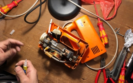 Auf einem Holztisch liegt ein orangefarbener Mixer. Er ist aufgeschraubt und man sieht das Innenleben des Mixers. Daneben liegt Werkzeug zum Reparieren des Ger&auml;ts.