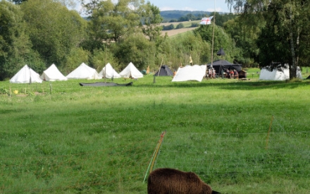 Zeltplatz mit Banner und verschiedenen Zelten (Jurte, Kohte, Wei&szlig;zelte). Im Vordergrund weidet ein Schaf.