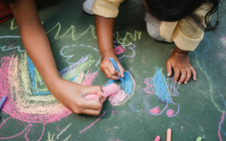 In Nahaufnahme sind Kinderh&auml;nde zu sehen, die mit roter und blauer Kreide Bilder auf einen dunkle Fl&auml;che malen.