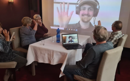 F&uuml;nf Senior:innen sitzen um einen Tisch, ein Mann hat einen Laptop vor sich stehen. Alle schauen auf eine Leinwand, auf der ein junger Mann mit Kopfh&ouml;rern zu sehen ist, der lacht und den Senior:innen zuwinkt.