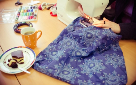 Auf einem Tisch steht eine N&auml;hmaschine, es sind die H&auml;nde einer Frau zu sehen, die mit einem lilafarbenem Stoff n&auml;ht. Daneben stehen eine Tasse Kaffee und ein St&uuml;ck Kuchen.