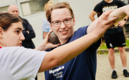 Zwei junge Frauen in Sportkleidung stehen beieinander. Die eine h&auml;lt eine Kugel in der Hand und die Hand mit der Kugel &uuml;ber der Schulter - so, als ob sie Kugelsto&szlig;en m&ouml;chte. Die andere Frau erkl&auml;rt etwas und zeigt die richtige Haltung f&uuml;rs Kugelsto&szlig;en.