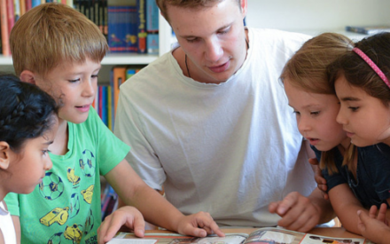 An einem Tisch sitzt ein junger Mann, rechtsl und links neben ihm Kinder im Grundschulalter. Alle schauen auf ein Buch, das vor ihnen liegt. Eins der Kinder zeigt mit dem Finger auf die Buchseite.