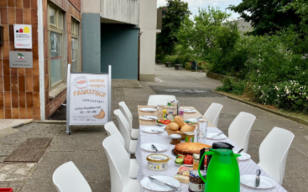 In einer Fu&szlig;g&auml;ngerzone steht ein gedeckter Fr&uuml;hst&uuml;ckstisch. Im Hintergrund ist ein Aufsteller zu sehen mit einem Plakat, auf dem der Schriftzug "Nachbarschaftsfr&uuml;hst&uuml;ck" steht.