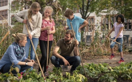 In einem Schulgarten knien zwei erwachsene Personen und sprechen miteinander. Drumehrum stehen mehrere Kinder mit Gartenwerkzeugen in der Hand, die arbeiten oder den Erwachsenen zuh&ouml;ren.