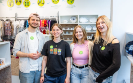 Vier junge Menschen stehen nebeneinander im Oxfam-Shop und l&auml;cheln in die Kamera. Sie tragen einen leuchtend gr&uuml;nen Button, der zeigt, dass Sie sich bei Oxfam engagieren.