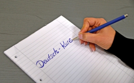 Auf einem Tisch liegt ein liniertes Blatt. Es ist eine Hand zu sehen, die einen blauen Stift h&auml;lt und schreibt. Auf dem Blatt steht der Schriftzug Deutsch-Kurs.