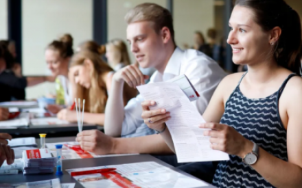 An einem Tisch sitzen eine junge Frau und ein junger Mann, vor sich Dokumente von der DKMS. Der Mann h&auml;lt einige Wattest&auml;bchen in der Hand, die Frau Infomaterialien. Beide sind offensichtlich mit Personen gegen&uuml;ber, die nicht auf dem Foto zu sehen sind, im Gespr&auml;ch. Im Hintergrund sind verschwommen viele weitere Menschen zu sehen.