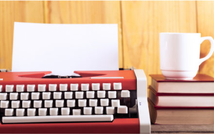 In Nahaufnahme ist eine rote Schreibmaschine mit wei&szlig;en Tasten zu sehen. Rechts daneben liegt ein Stapel B&uuml;cher auf der eine wei&szlig;e Tasse steht.