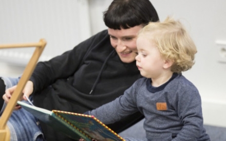 Eine junge Frau und ein Kleinkind liegen und sitzen gemeinsam auf dem Fu&szlig;boden. Beide schauen in ein Kinderbuch. Das Kind zeigt auf etwas in dem Buch.