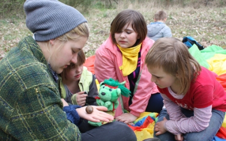 Auf einer Decke auf dem Boden sitzen 3 Kinder und eine jugendliche Betreuerin. Die Betreuerin zeigt den Kindern das Geh&auml;use einer Weinbergschnecke, dass auf ihrer Handfl&auml;che liegt.