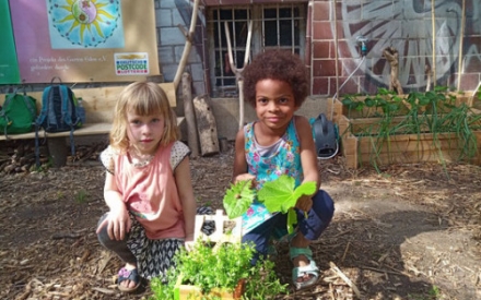 Zwei Kinder sitzen auf der Erde. Im Hintergrund ist eine bunt bemalte Mauer. Vor den Kindern steht ein Holgef&auml;&szlig; mit Pflanzen. Eines der Kinder h&auml;lt zwei gro&szlig;e Bl&auml;tter in den H&auml;nden.