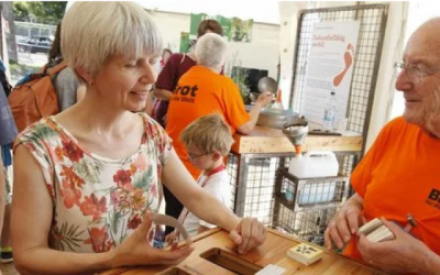 Ein Mann und eine Frau stehen sich gegen&uuml;ber. Die eine Person tr&auml;gt ein leuchtend orangefarbenes T-Shirt mit dem Schriftzug "Brot f&uuml;r die Welt". Beide sind im Gespr&auml;ch und schauen nach unten auf eine Art Spiel mit Karten.