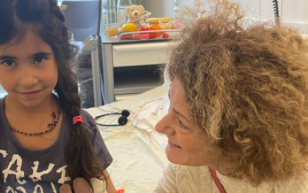 Ein kleines M&auml;dchen sitzt auf einem Krankenhausbett. Es hat einen verbundenen Arm, h&auml;lt ein Kuscheltier in der Hand und l&auml;chelt sch&uuml;chtern in die Kamera. Neben ihr sitzt eine Frau, sie wendet sich dem Kind zu und l&auml;chelt es an.