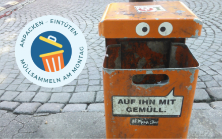 Ein gro&szlig;er orangefarbener M&uuml;lleimer aus Metall steht an einer gepflasterten Stra&szlig;e. Auf der oberen Frontseite des M&uuml;lleimers sind zwei gro&szlig;e Augen aufgeklebt. Auf dem unteren Teil ist ein Aufkleber mit der Aufschrift "Auf ihn mit Gem&uuml;ll"