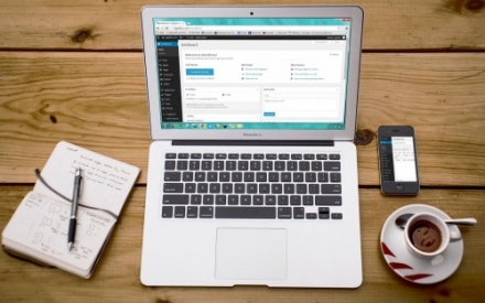 auf einer Holztischplatte steht ein ge&ouml;ffneter Laptop. AUf dem Bildschirm ist eine Webseite zu sehen. Neben dem Computer sind eine Tasse Kaffee, ein Smarthphone und ein Notizblock samt Stift