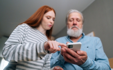 Nahaufnahme eines &auml;lteren Mannes mit Vollbart, der gemeinsam mit einer jungen Frau auf sein Mobiltelefon schau und etwas erkl&auml;rt bekommt.