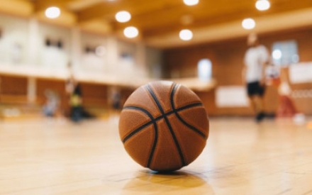 Ein Baskettball liegt auf dem Holzboden einer Turnhalle, ganz im Vordergrund, in der rechten Ecke des Fotos. Der Hintergrund der Halle ist nur unscharf zu erkennen, Menschen und Lichter sind nur schemenhaft zu sehen.