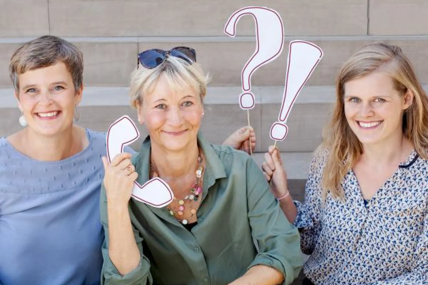 3 l&auml;chelnde Frauen sitzen auf einer Treppe und halten Schildchen hoch