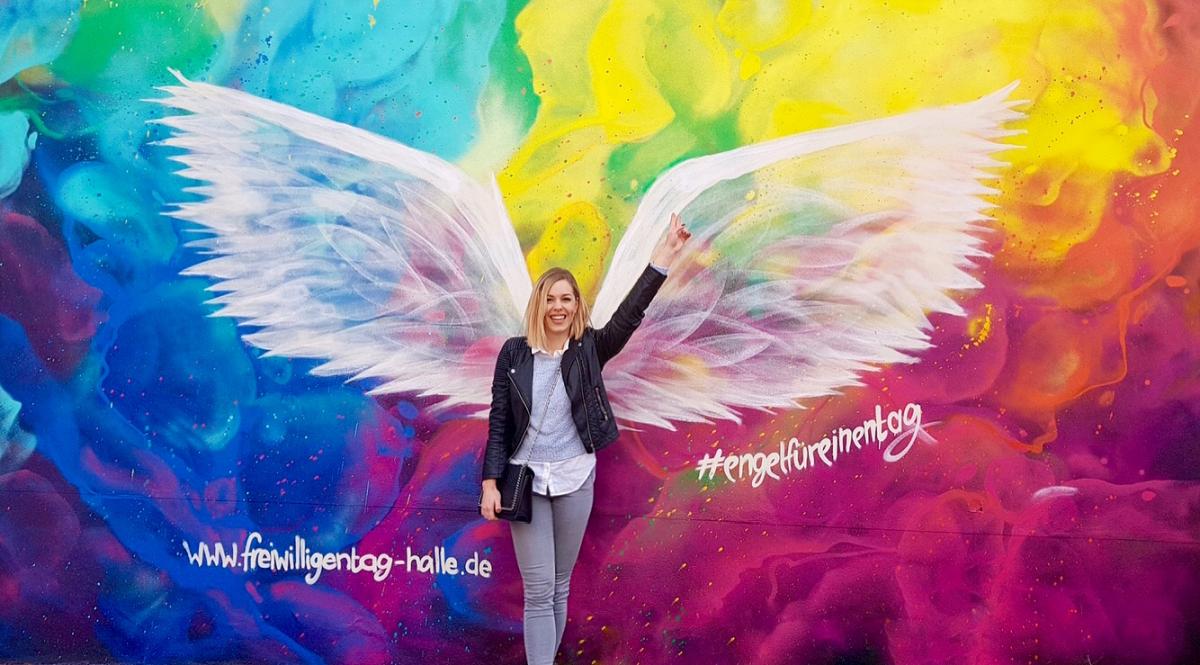 eine junge Frau steht vor einer bunten Wand, an die weiße Flügel gemalt sind