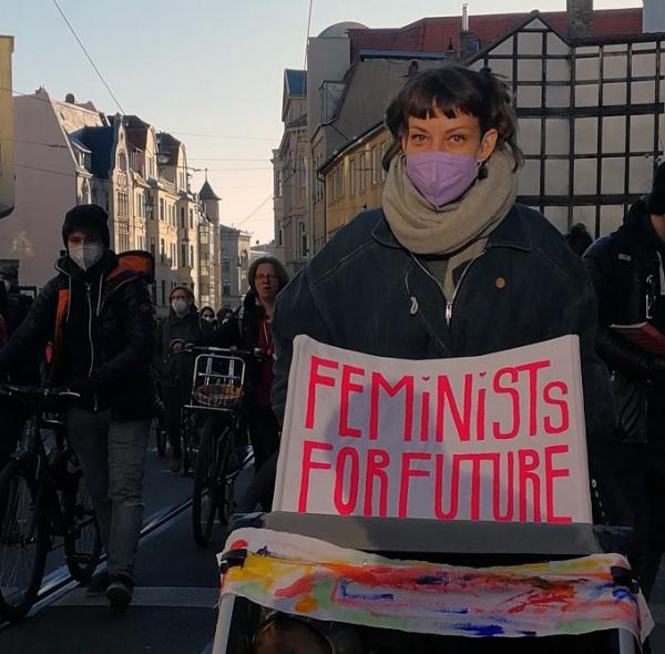 Zu sehen ist eine junge Frau auf einer Demo. Sie trägt ein Schild, auf dem "Feminists for Future" steht.