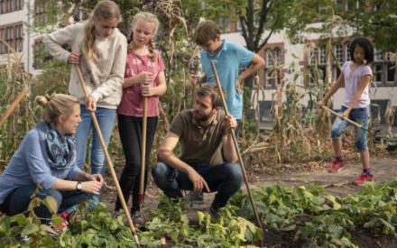 In einem Schulgarten knien zwei erwachsene Personen und sprechen miteinander. Drumehrum stehen mehrere Kinder mit Gartenwerkzeugen in der Hand, die arbeiten oder den Erwachsenen zuhören.